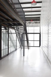 Industrial warehouse interior with a metal staircase, mezzanine level, large grid windows, and bright white flooring.