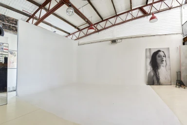 Empty white photo studio with high truss ceiling, red pendant lights, and a large portrait canvas leaning against the wall.