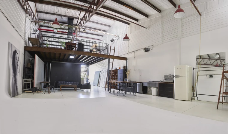 Spacious industrial photo studio in Melbourne with white walls, mezzanine level, red pendant lamps, and concrete floor.