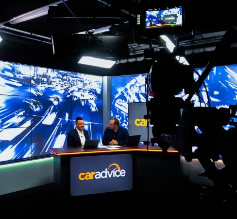 Two adult males on a television news set in front of monitors. A large professional camera is visible in the foreground.