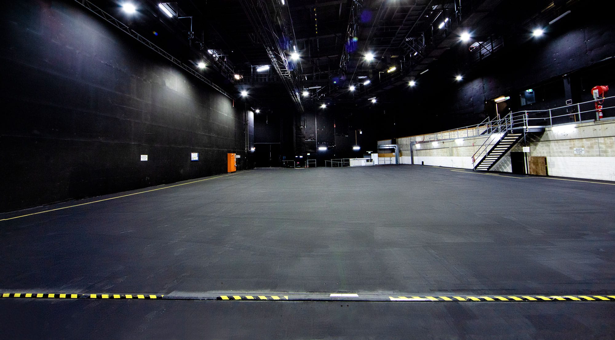 Wide shot of a large, empty photo studio with dark walls, a grey floor, and overhead studio lighting, suitable for photography or video production.