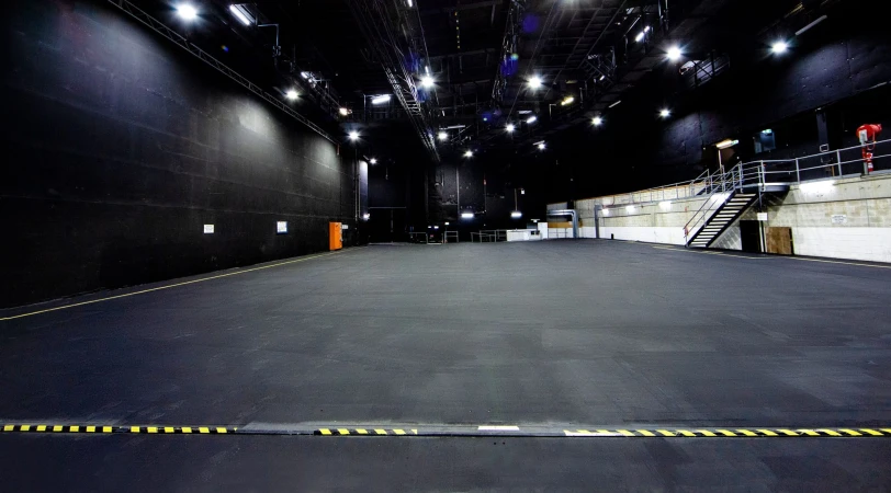 Wide shot of a large, empty photo studio with dark walls, a grey floor, and overhead studio lighting, suitable for photography or video production.