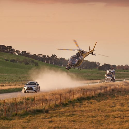 Aerial shot of silver car and helicopter kicking up dust on rural road with green hills, trees, and semi-truck in background at sunset.