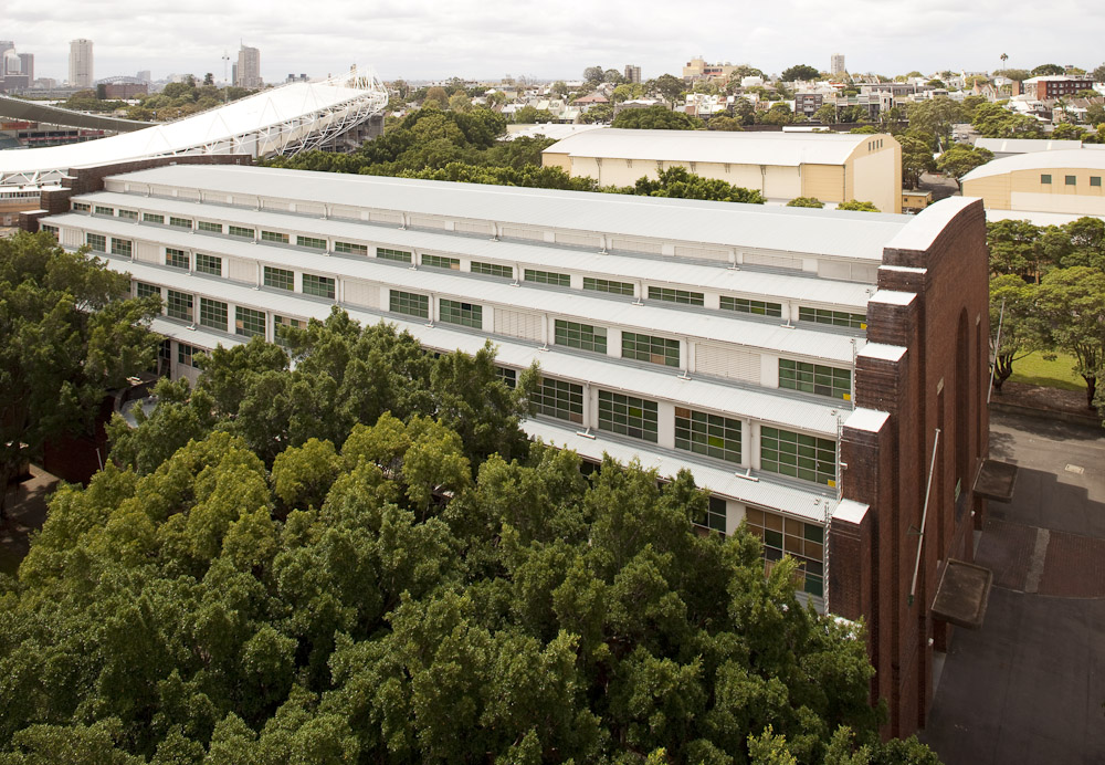 Overhead shot of a modern brick building with numerous windows, surrounded by green trees and other buildings in the background.