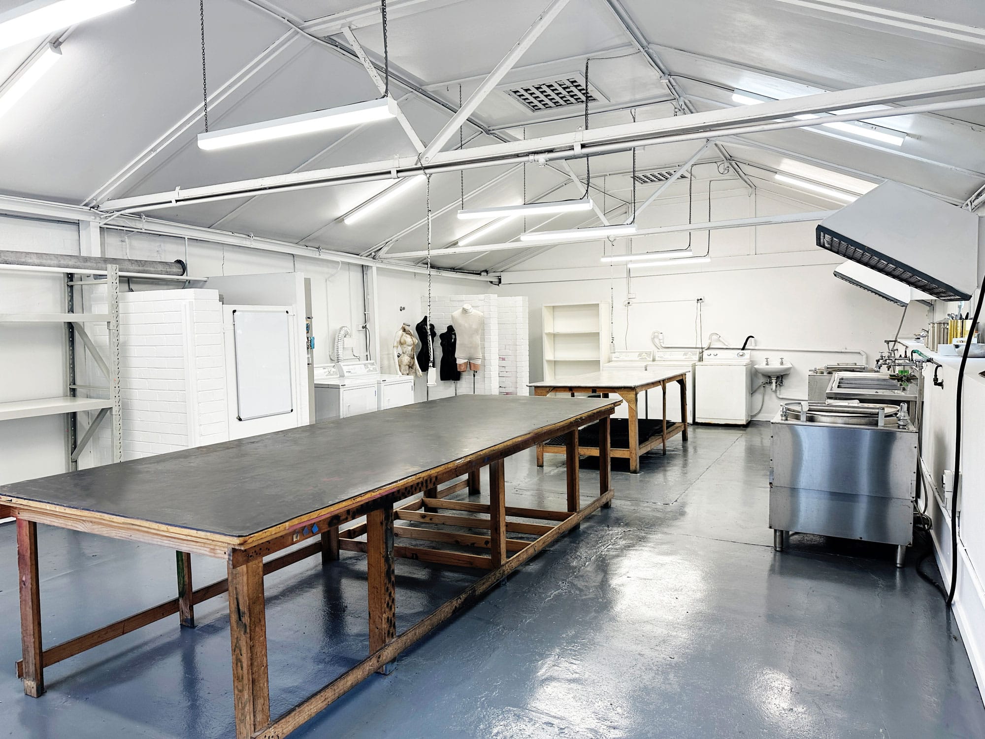 Spacious white industrial setting with large wooden tables, stainless steel equipment, and overhead lighting in a clean, organized layout.