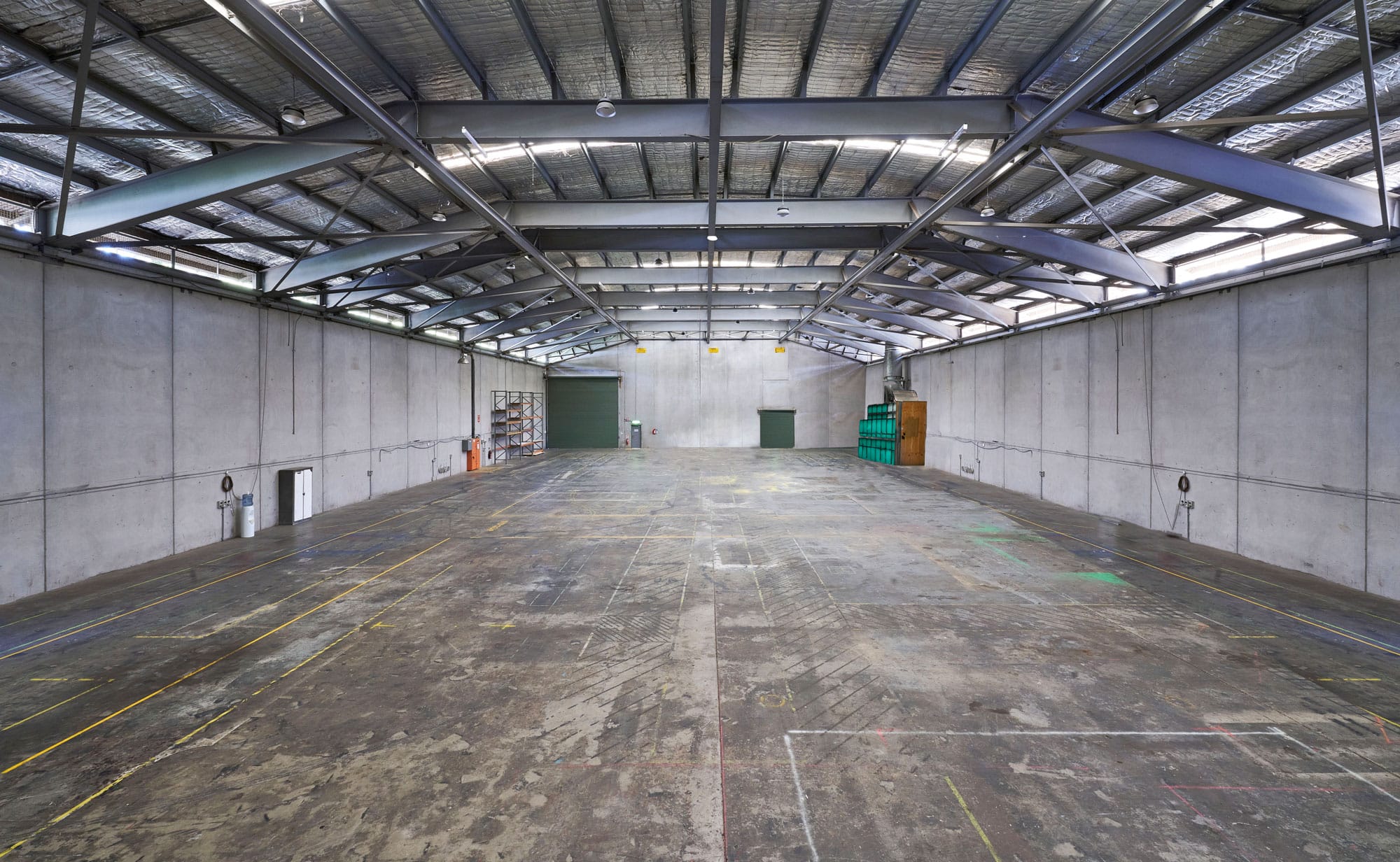 Large empty warehouse interior with concrete floor, metal beams, and open space; natural and artificial lighting creates stark industrial scene.