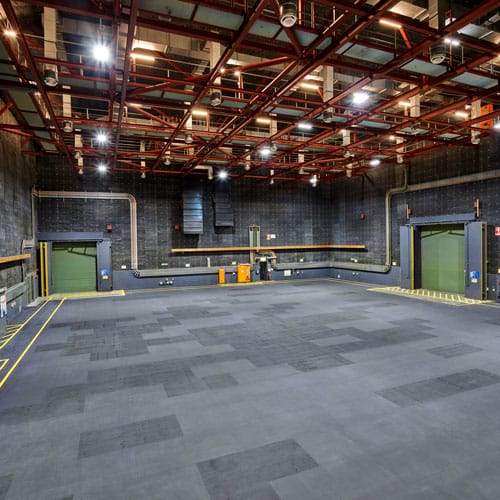Wide shot of a large empty studio space featuring dark gray carpet, gray cinderblock walls, and a red iron framework ceiling.