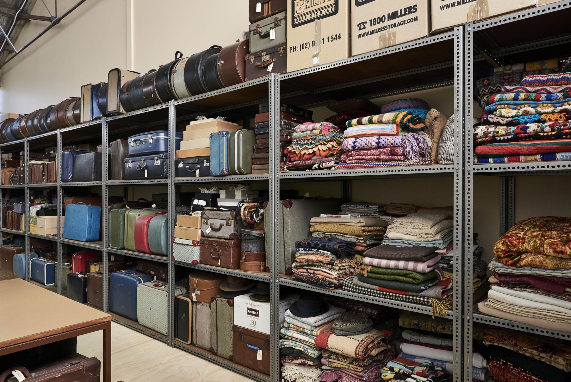 Shelves filled with vintage suitcases, boxes and folded blankets in a storage room. The objects are arranged neatly.