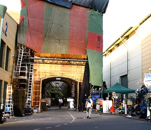 Film set with large tarps covering structures, scaffolding, lighting equipment, and people working on the lot in daylight.