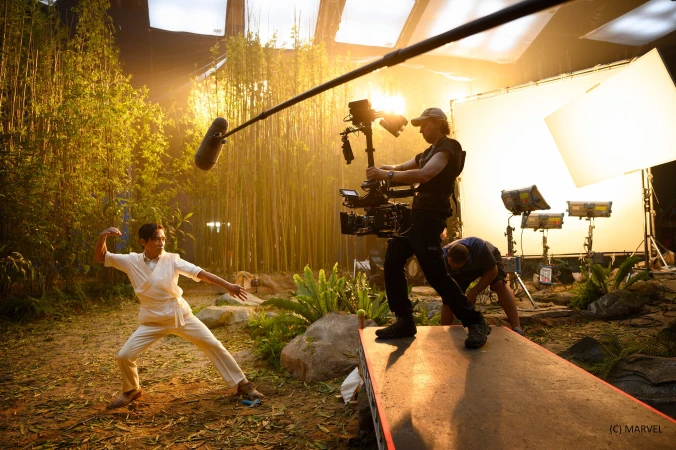 On a movie set, a male actor in a white outfit performs a martial arts stance while a cameraman films the scene surrounded by bamboo.