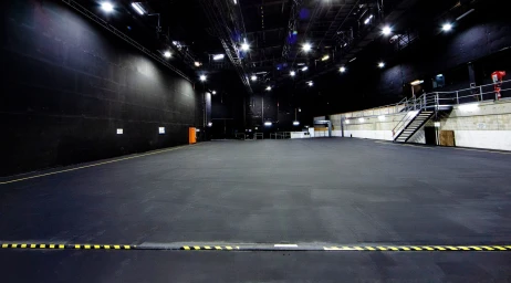 Wide empty industrial film soundstage in Sydney with dark walls, large dark grey floor, overhead studio grid lighting, and elevated catwalk
