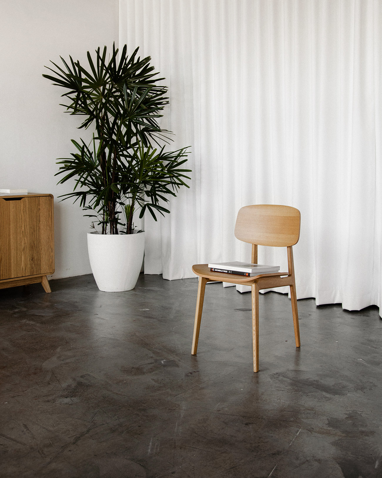 Interior design featuring a wooden chair with a book, a potted palm, and a wooden cabinet against a white curtain backdrop.