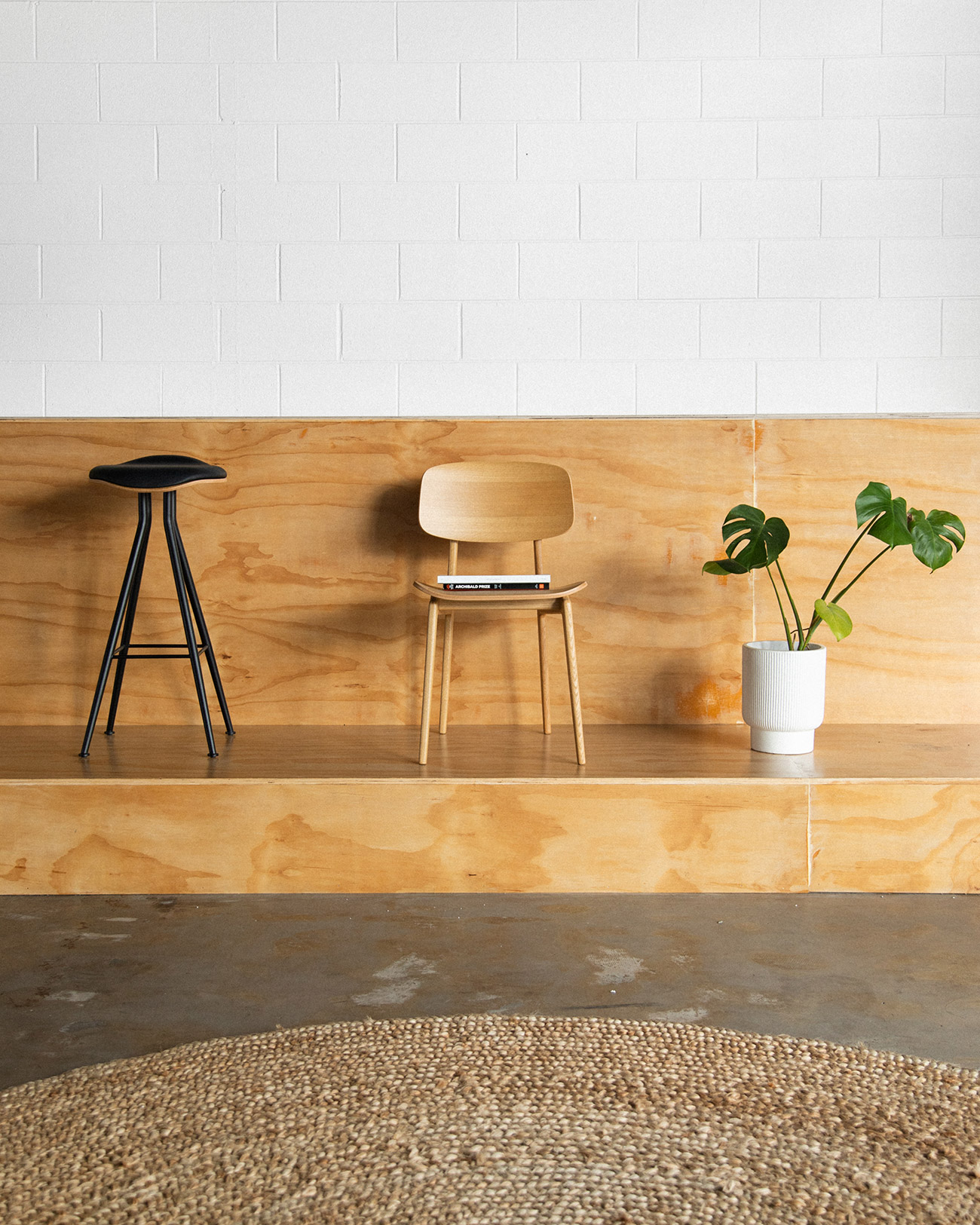 Interior shot of a light wood chair, black bar stool, and potted plant against a white brick and wood paneled wall on a concrete floor.