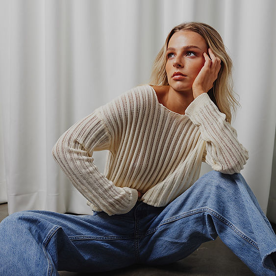 A young adult woman with blonde hair is seen indoors sitting and looking up, wearing a beige ribbed top and jeans in a studio setting.