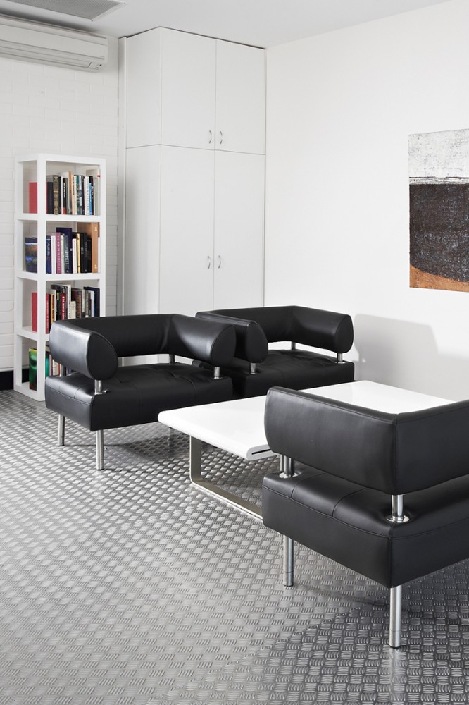 Indoor waiting area with three modern black leather chairs and a white table on a textured gray floor, alongside a bookshelf and storage cabinet.