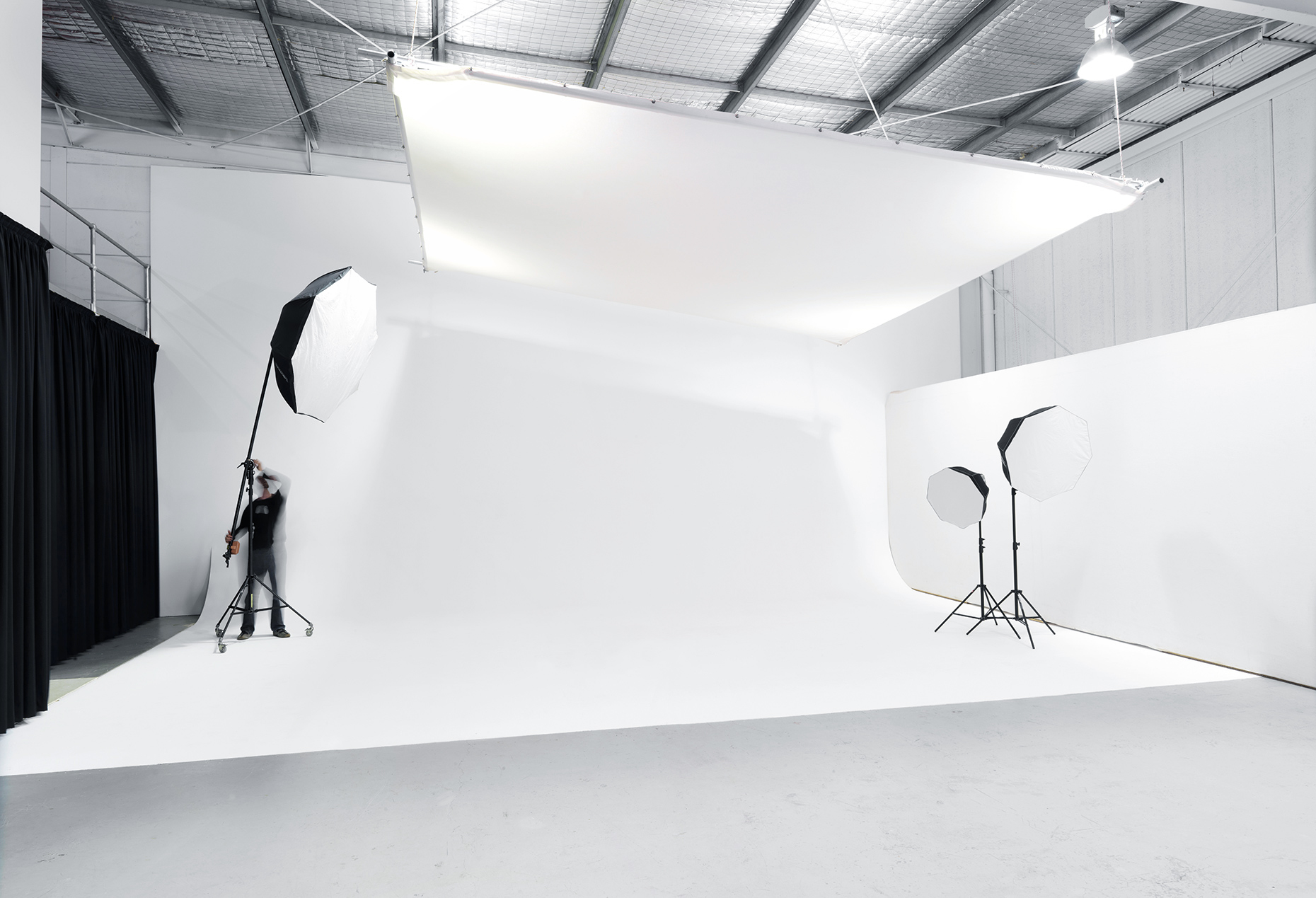 Wide shot of a spacious photo studio with a white backdrop, softbox lighting, and black curtains, ready for a professional photoshoot.