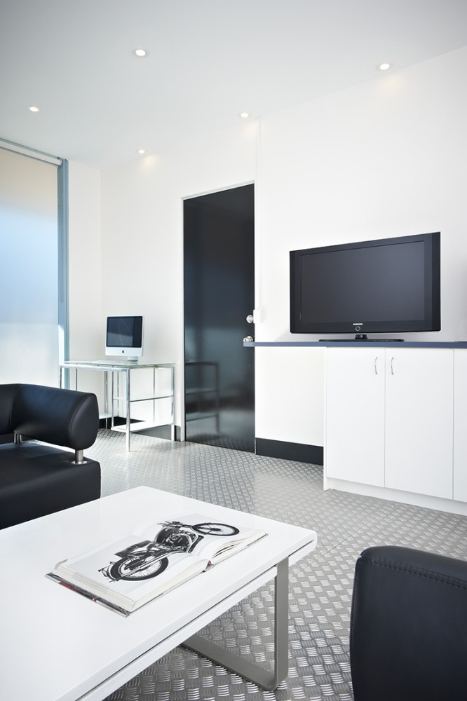 Bright, modern interior with black sofa, white table with a motorcycle magazine, flat-screen TV, computer, and silver diamond-plate flooring.
