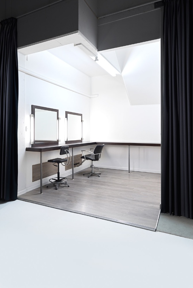 Photo studio interior with dark curtains, two vanities with mirrors and rolling chairs, and a gray wood floor. Bright, neutral color palette.