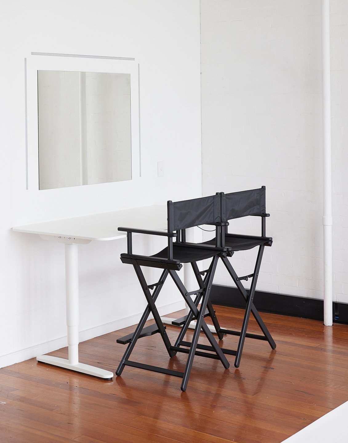 Photo studio setup with two black director's chairs in front of a white table and mirror, on a wooden floor in a bright room.