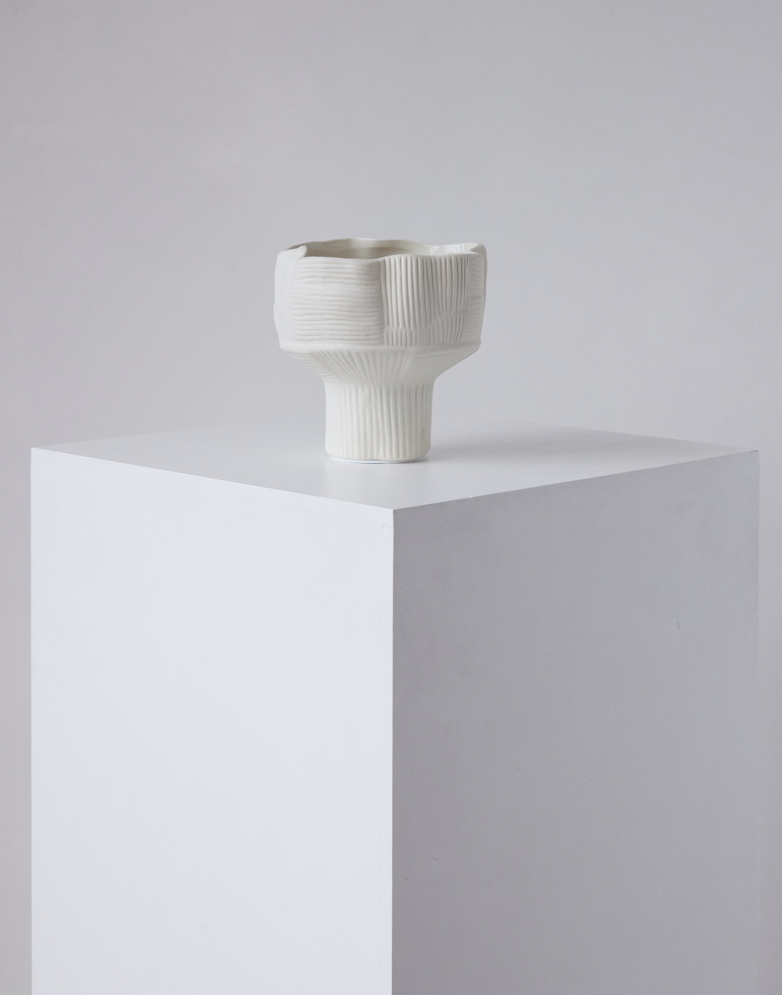 Minimalist white ceramic vase with textured vertical lines, displayed on a white cubic pedestal against a soft grey background in studio lighting.