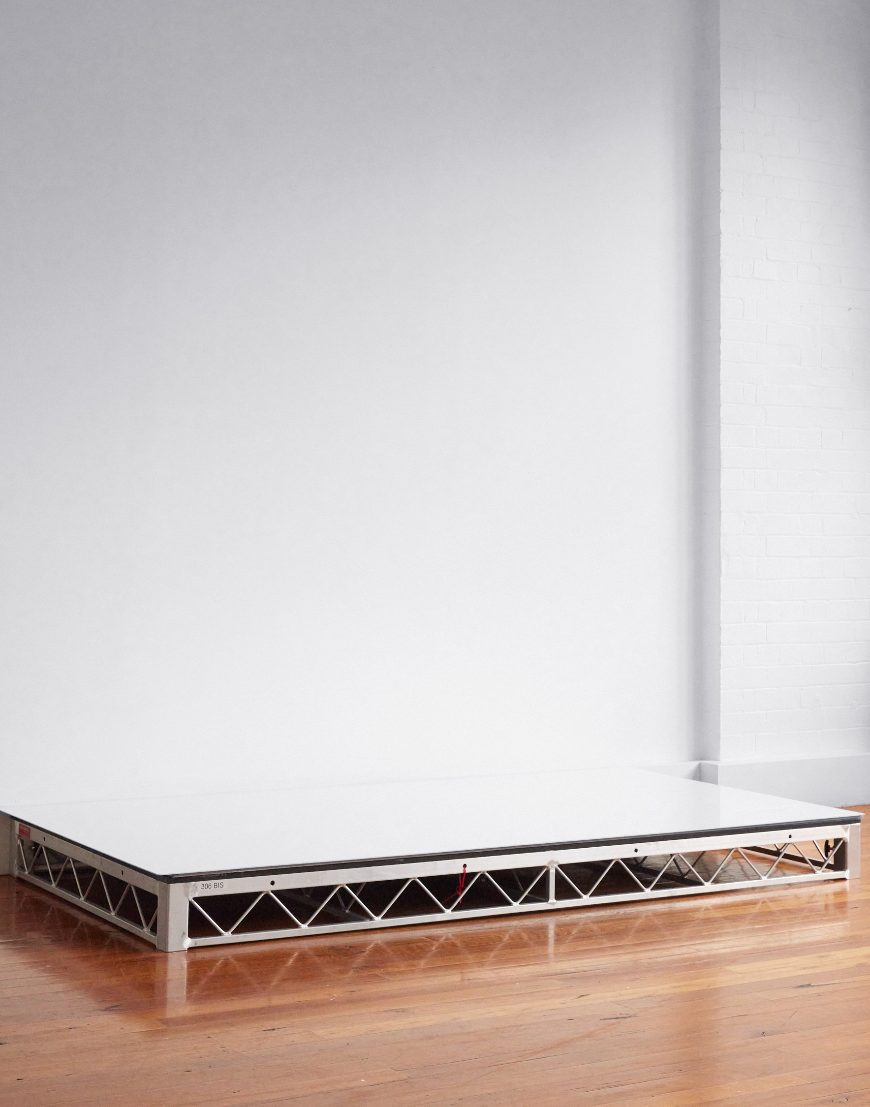 Minimalist photography of an elevated white platform with metal supports against a white wall and hardwood flooring in natural light.