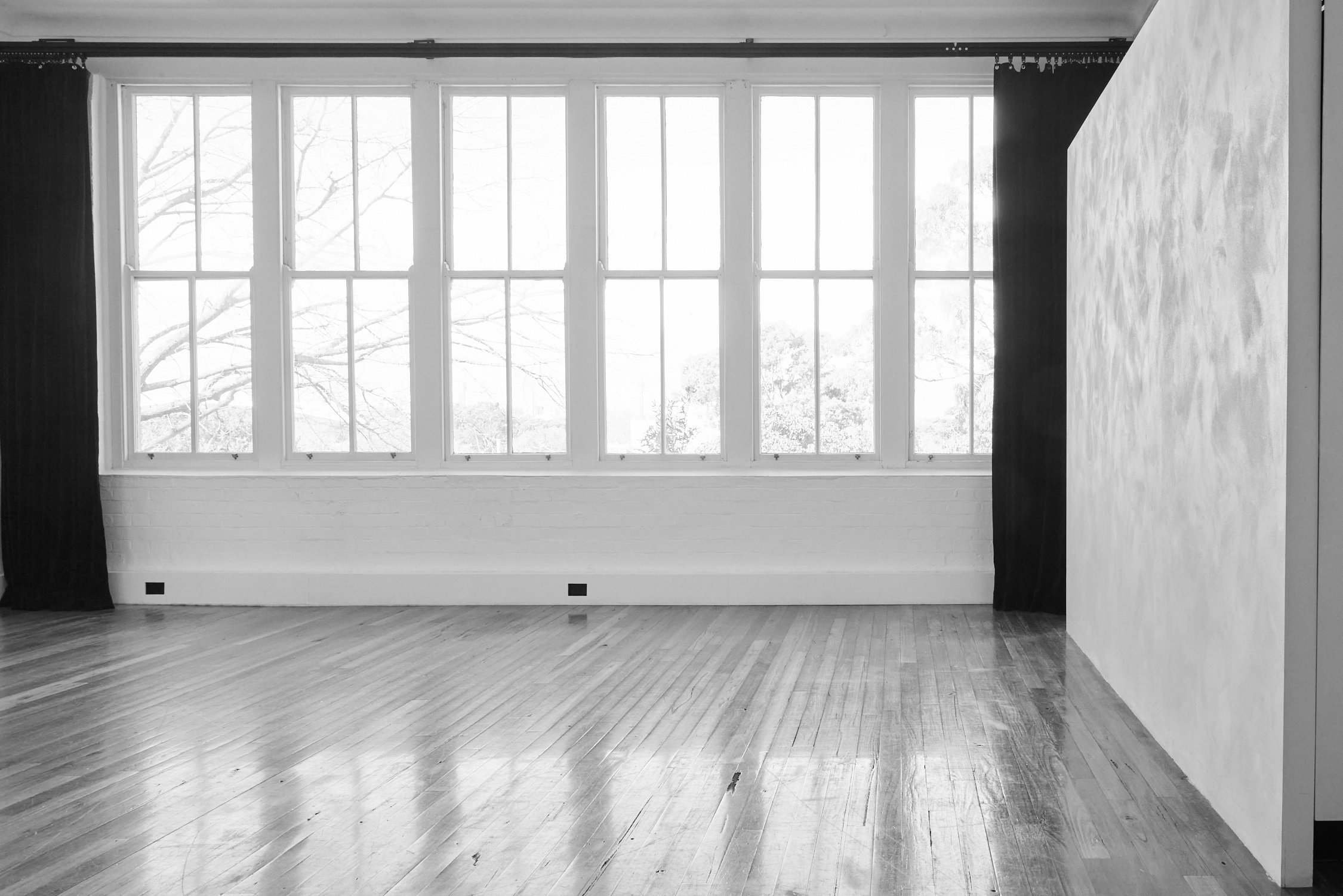 Bright, empty photo studio with large windows, white walls, and hardwood floor, featuring a textured screen and dark curtains.