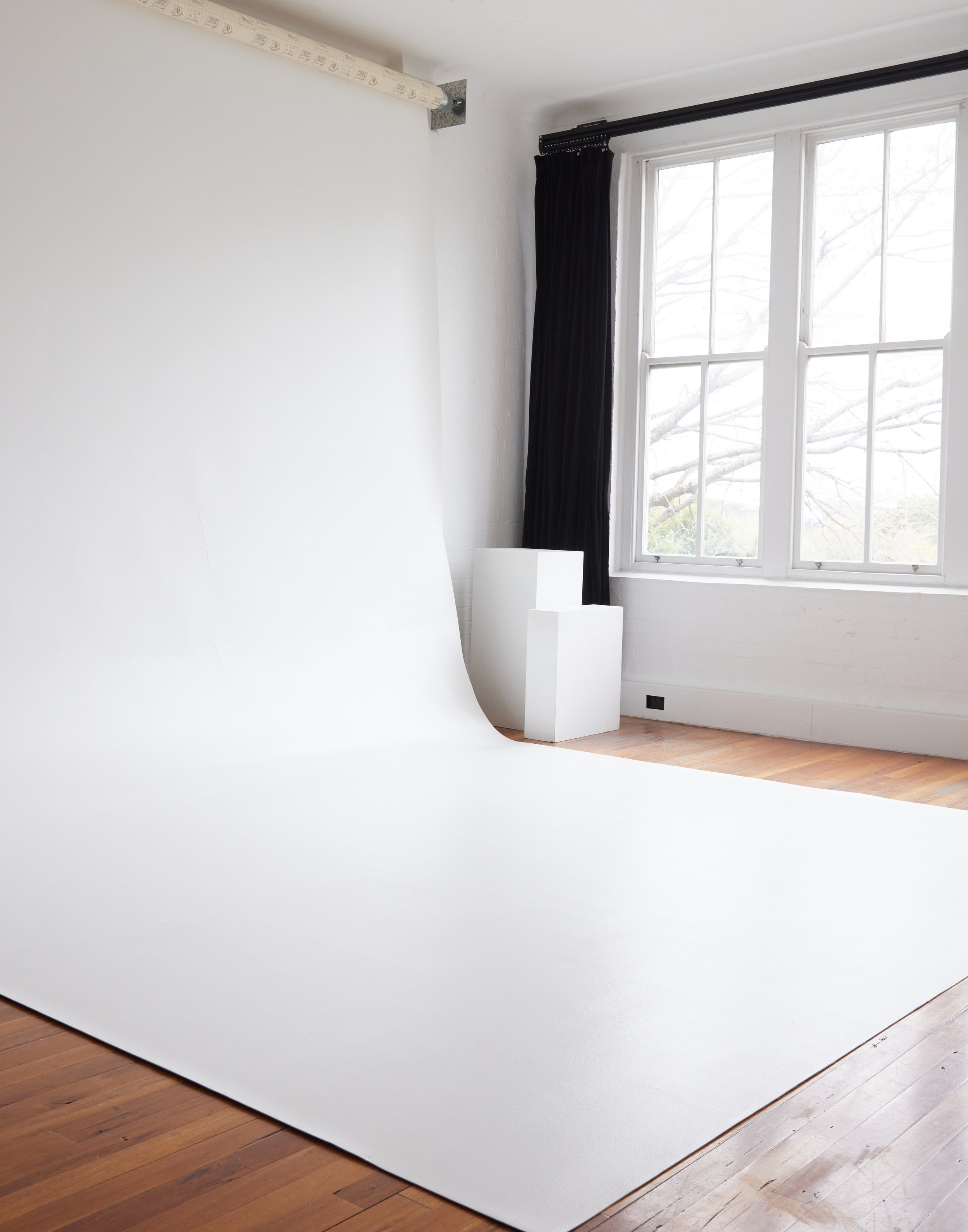 Photo studio with white seamless backdrop, white geometric pedestals, wooden floor, natural light from a window with black curtains.