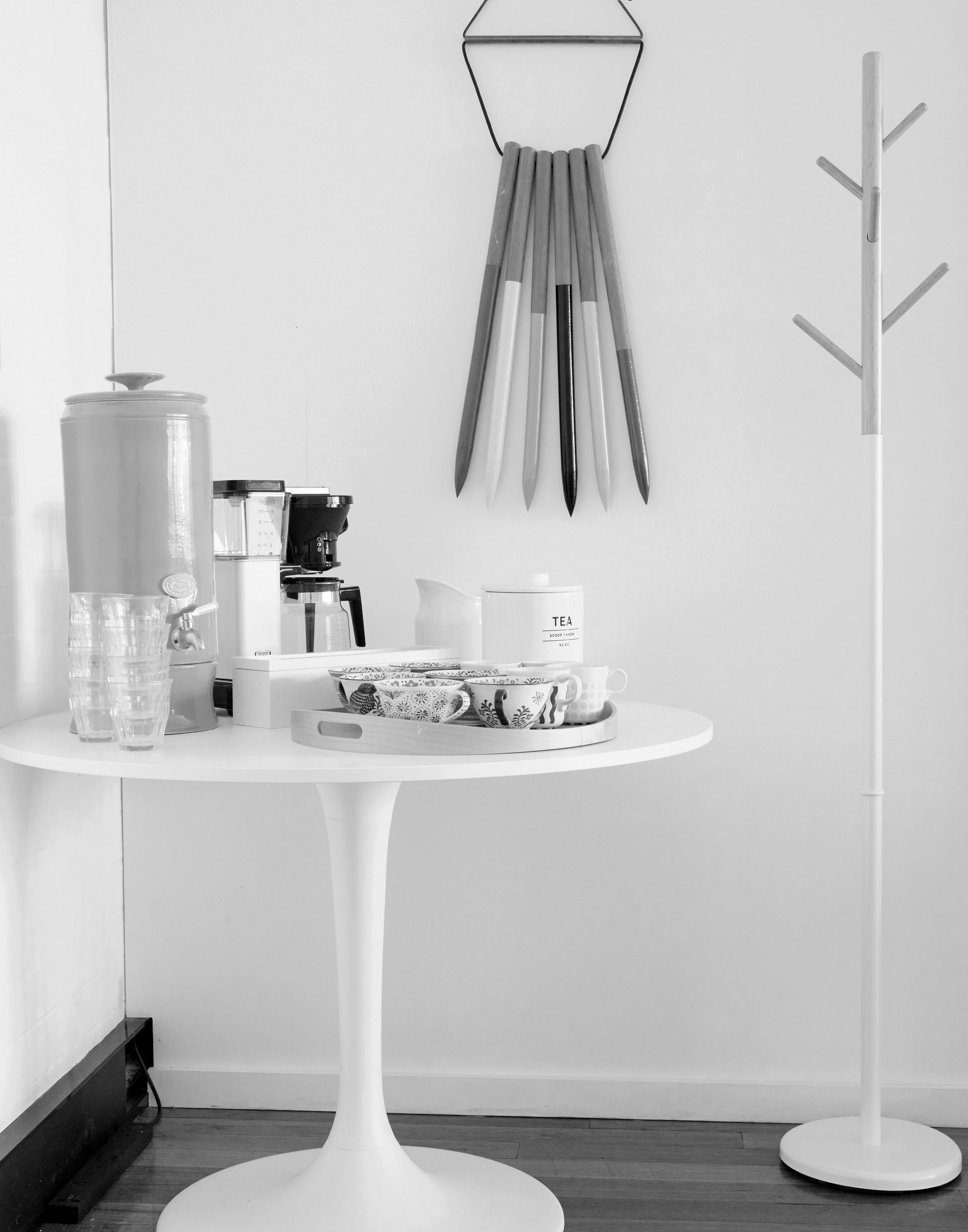 Black and white image of tea cups on a round table with a coffee maker and tea pot, with a coat rack and wall decoration in background.