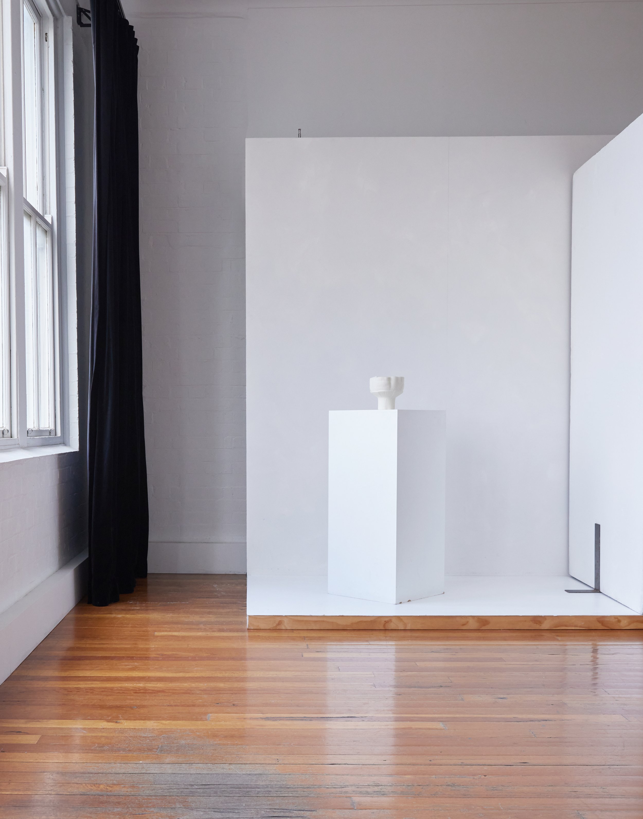 Minimalist interior shot of white plinth with a white sculptural vase, hardwood floors, white walls, and a window with a black curtain.