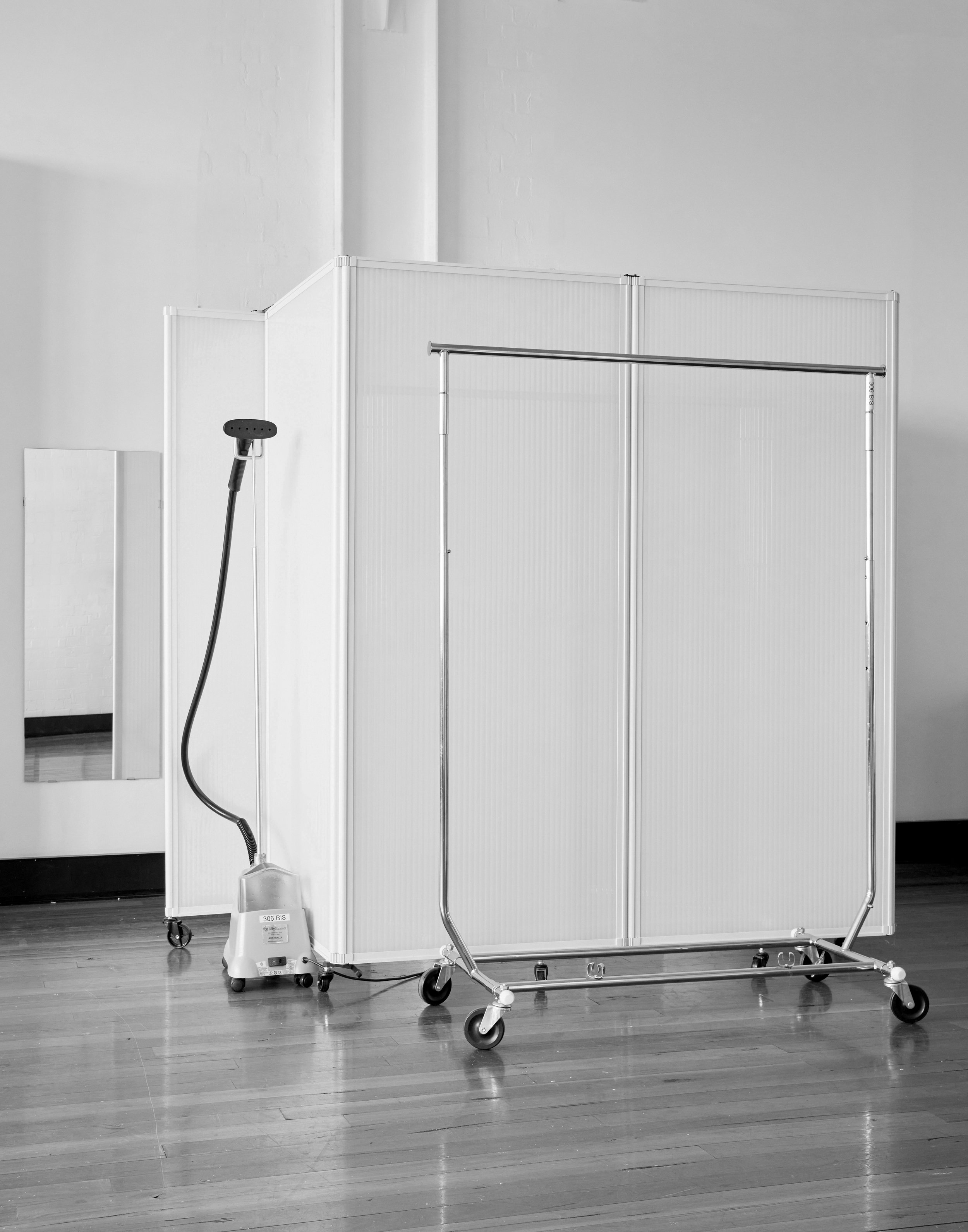 Image of clothing rack, garment steamer, and dressing room divider against a white wall, set on a wood floor in a photo studio.
