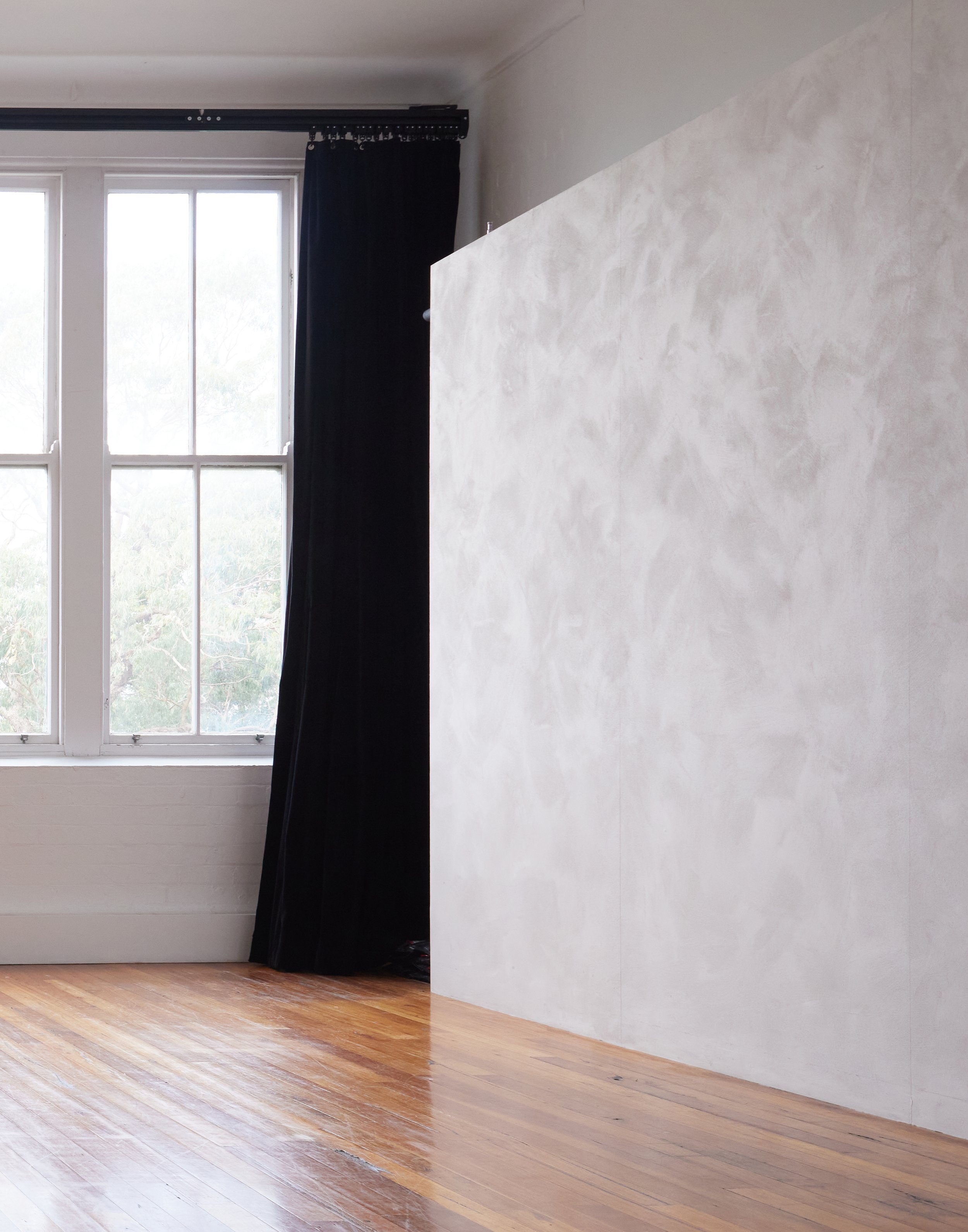 Interior view of an empty photo studio with a large window with black curtains, wooden floor, and grey textured backdrop.