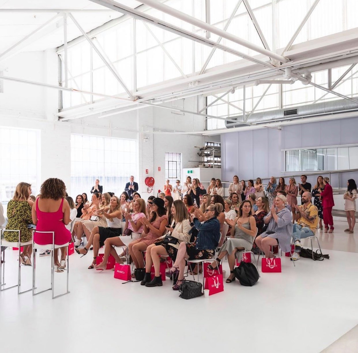 Audience of women and a few men clapping in a bright, white event space, with a woman in pink speaking to them from a chair.