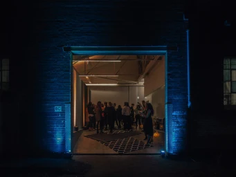 Exterior night view looking through an open industrial doorway at a gathering of people inside a lit Sydney event space.