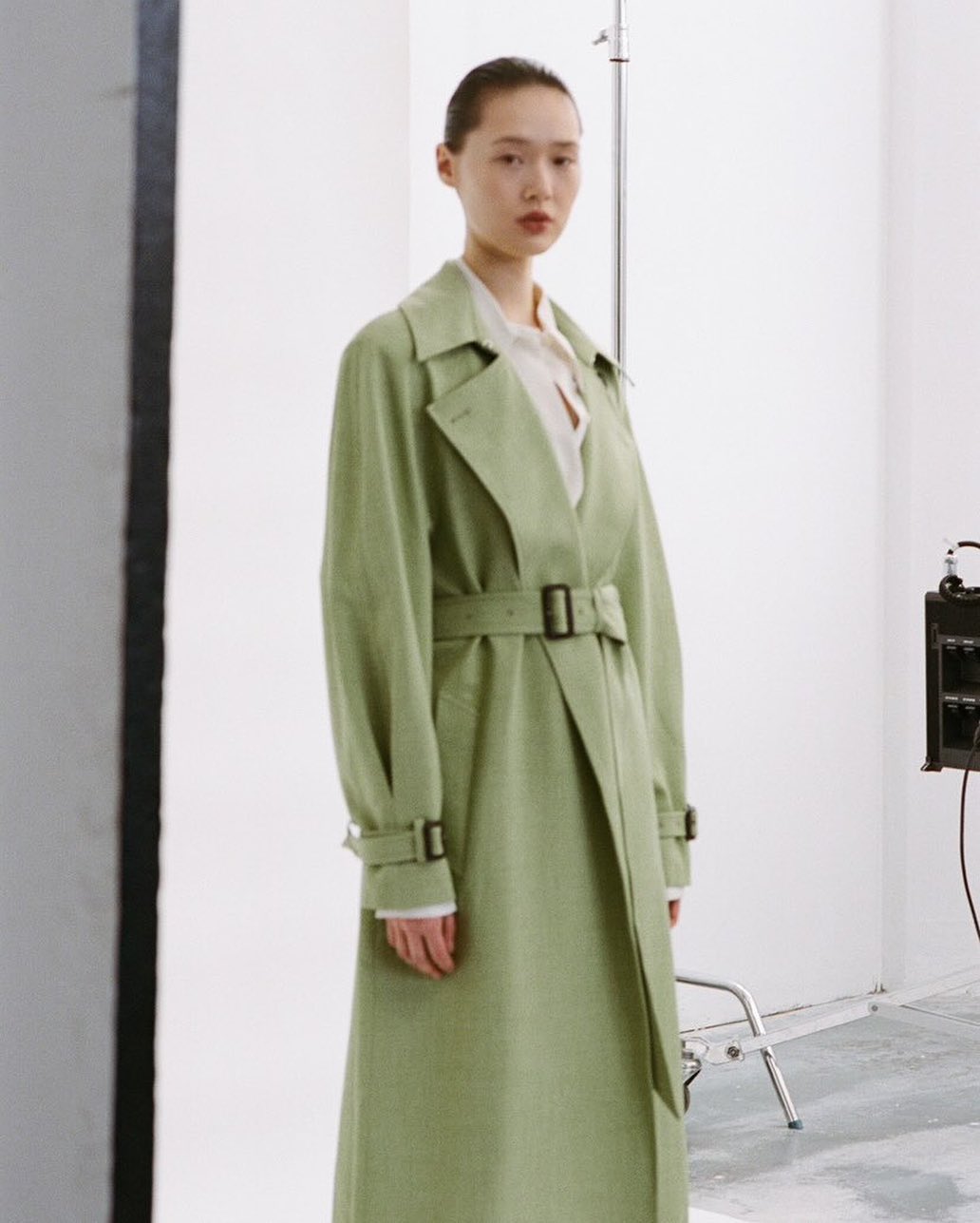 Elegant adult woman in a light-green trench coat stands in a studio. The coat is belted and she wears a white collared shirt underneath.