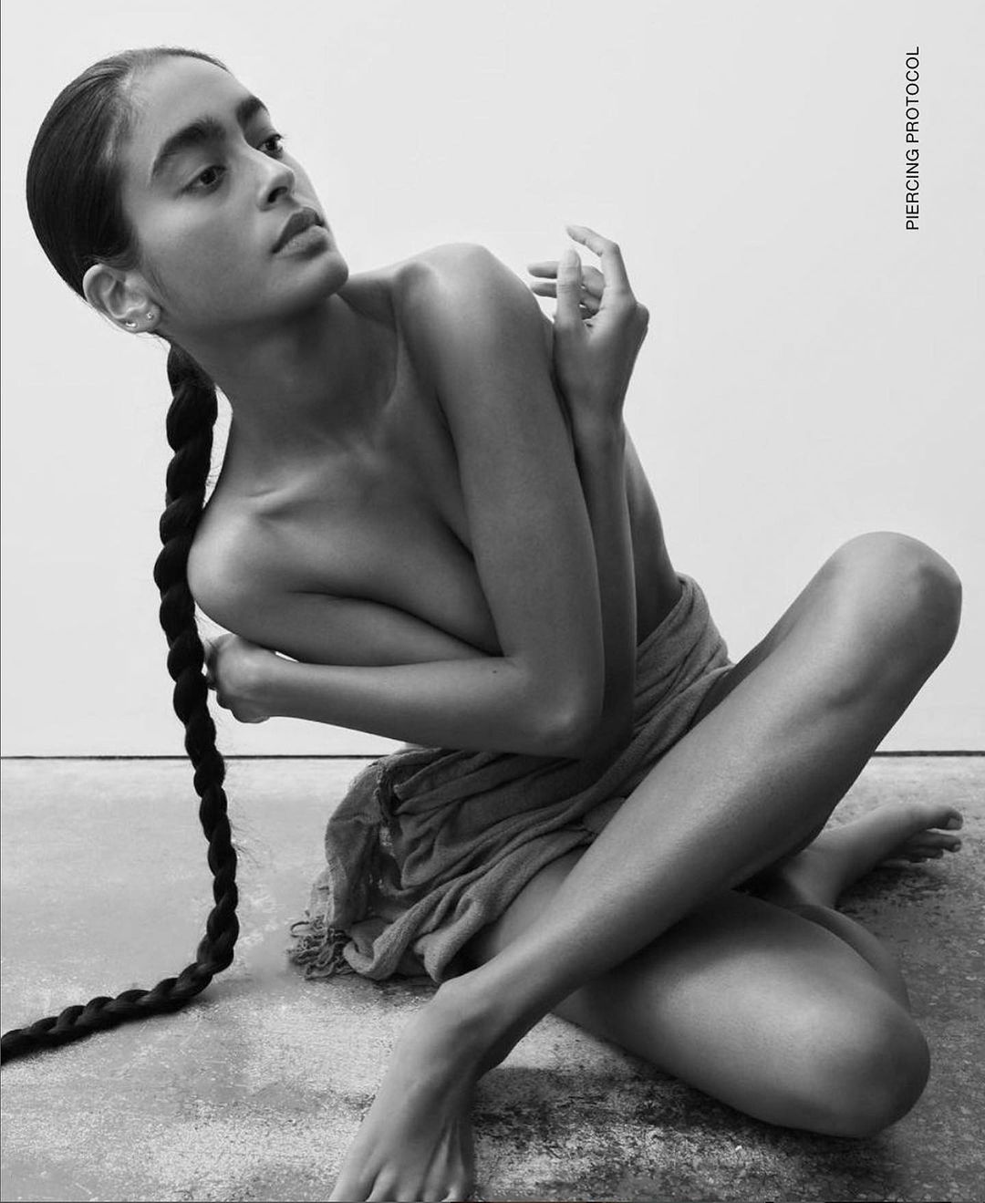 Black and white portrait of an adult female with long braid, arms and legs crossed, indoors against a neutral background.
