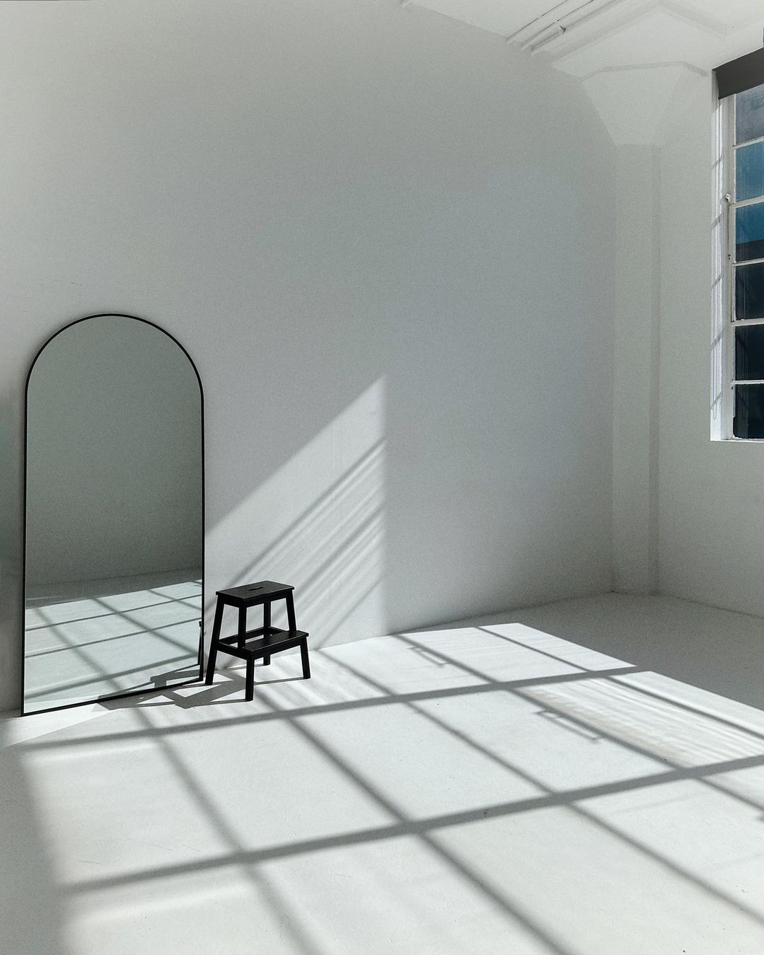 Minimalist studio with rounded black mirror, black step stool, and window light casting graphic shadows on the white walls and floor.