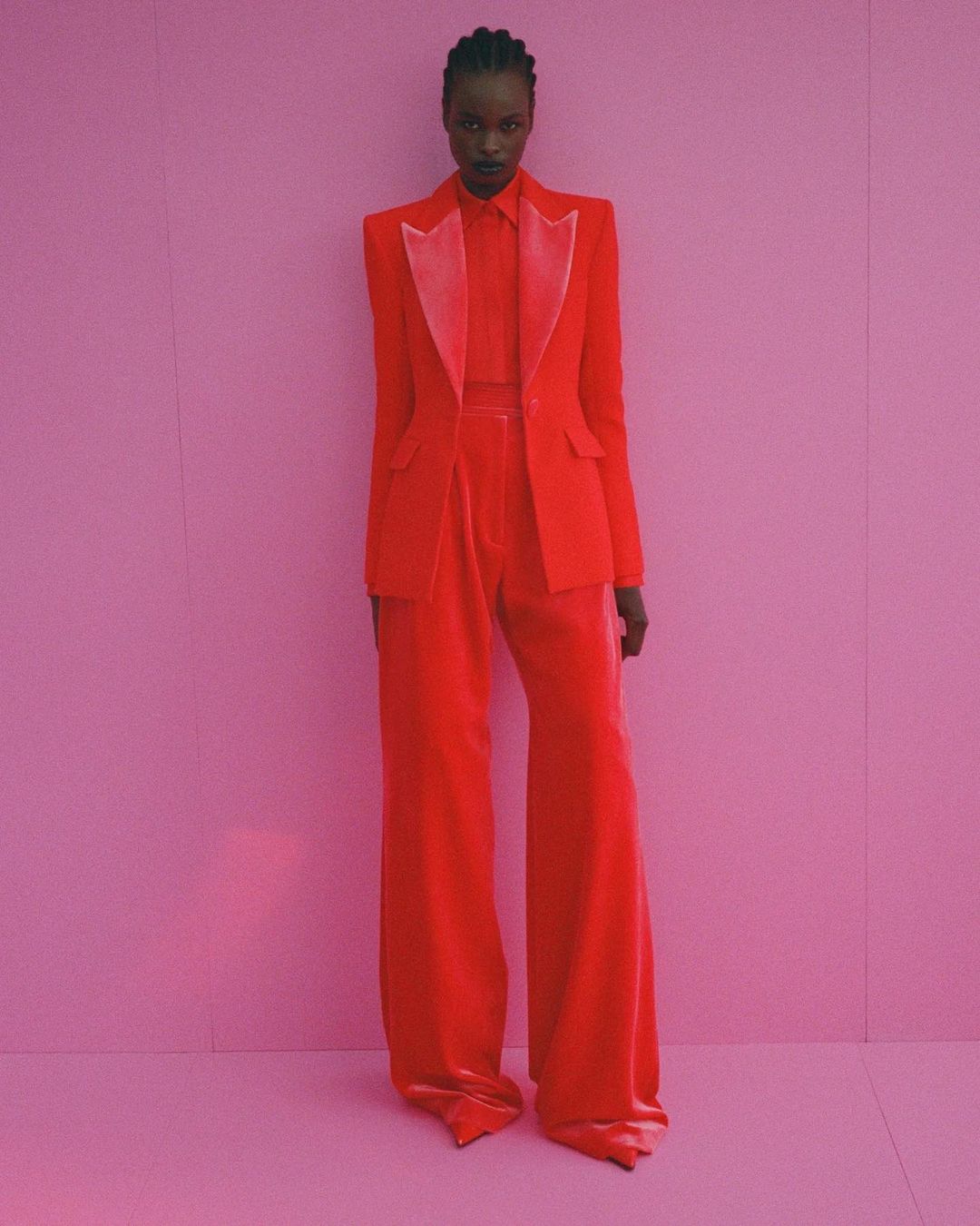 Elegant Black woman in a striking red suit stands against a vibrant pink backdrop, showcasing a modern fashion statement.