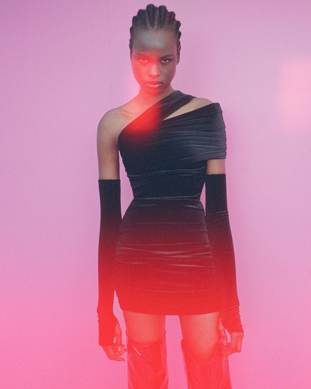 Young African woman in a black, form-fitting dress with sleeves and boots, posing against a pink background with red lighting effects.