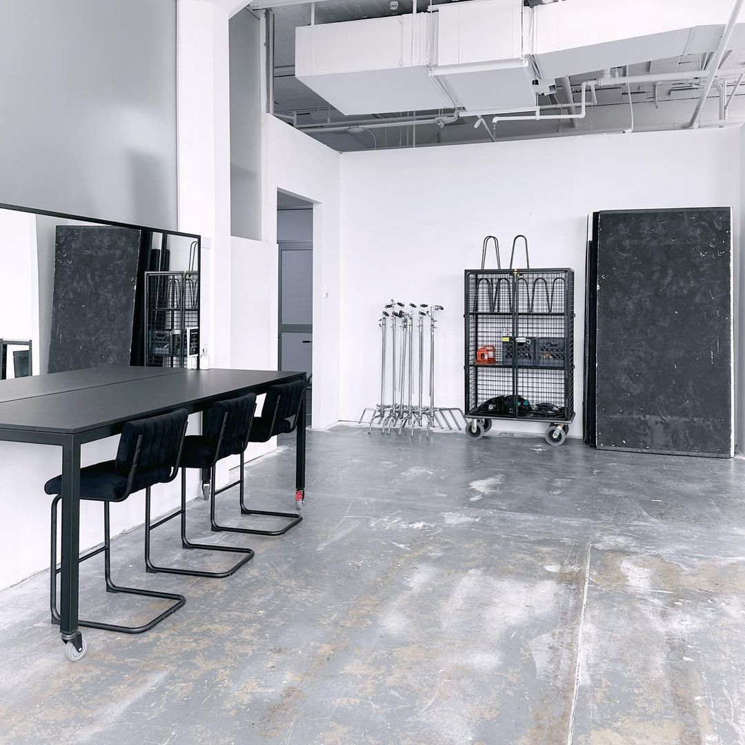 An open, bright photo studio with a black table, chairs, lighting equipment, and concrete floors, offering a versatile space for photography.