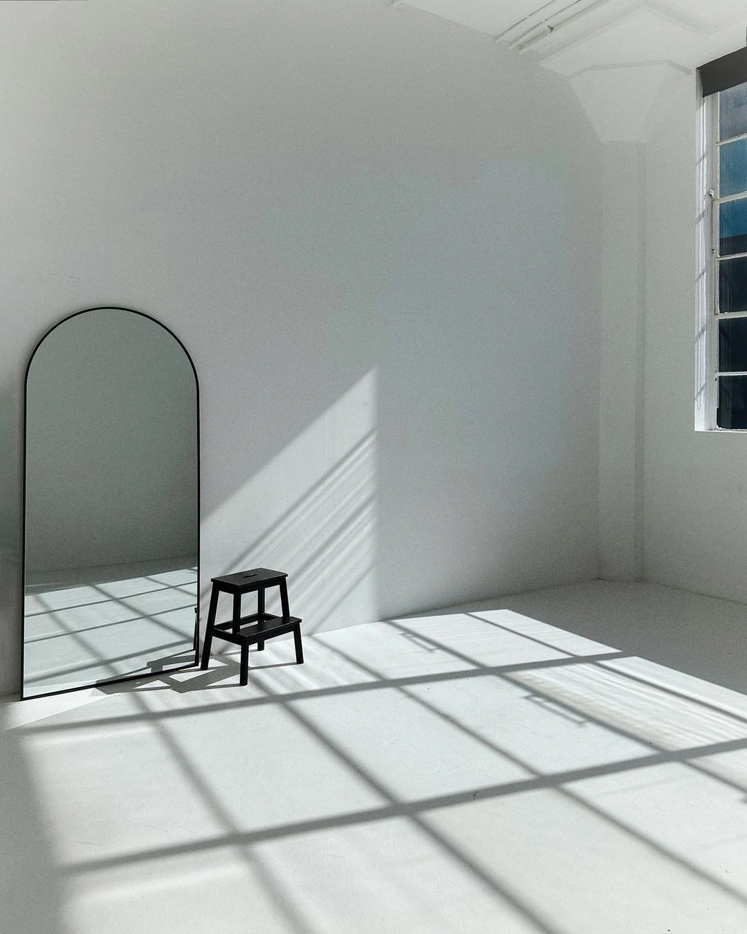 An arched floor mirror and a small black stool inside a bright white photography studio with diagonal window light casting shadows.