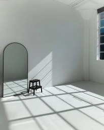 An arched floor mirror and a small black stool inside a bright white photography studio with diagonal window light casting shadows.