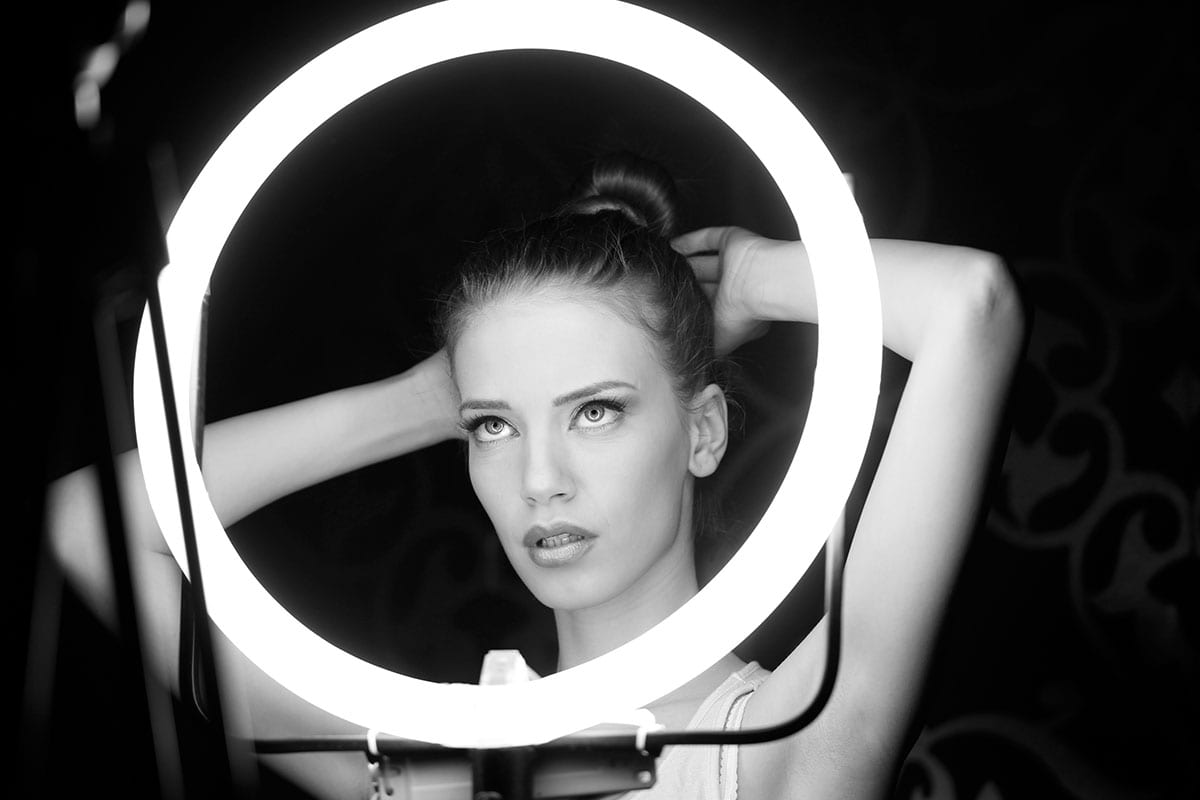 Black and white portrait of a fair-skinned woman with her hands behind her head, illuminated by a ring light, set against a dark backdrop.