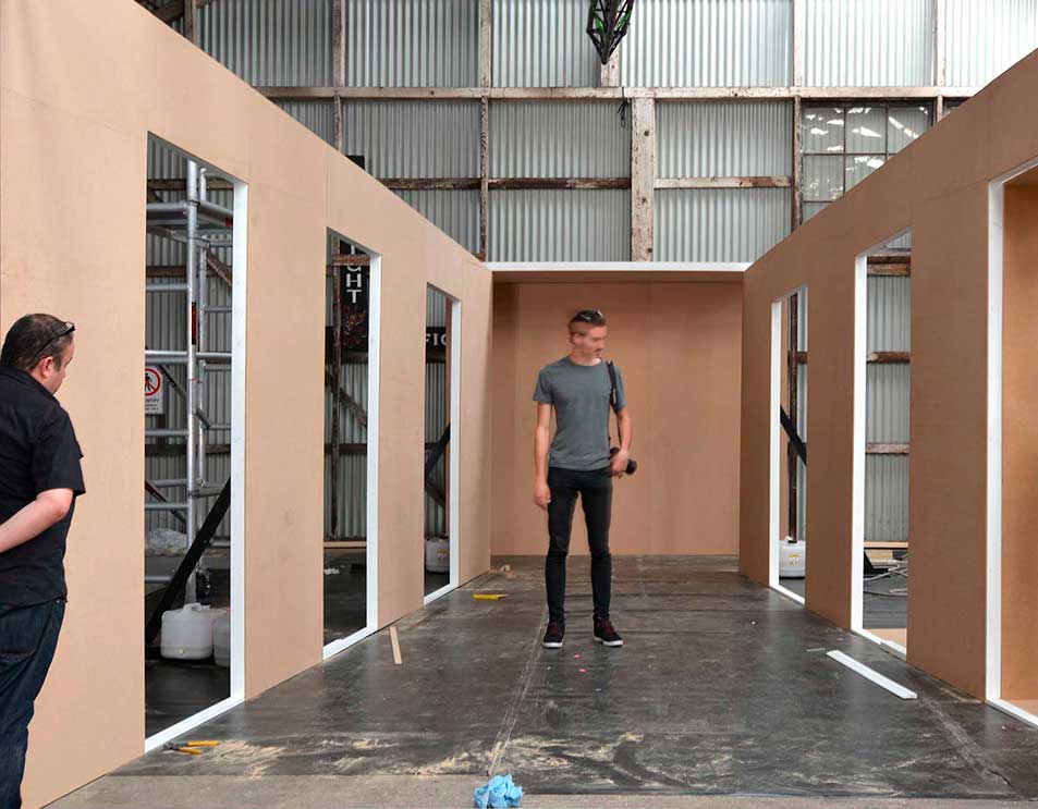 Two men stand in an industrial setting next to walls with door frames. One holds a camera, while metal beams support the ceiling.
