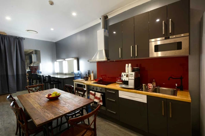 Modern kitchen and makeup room with dining table. Gray cabinets, red backsplash, and vanities with illuminated mirrors.