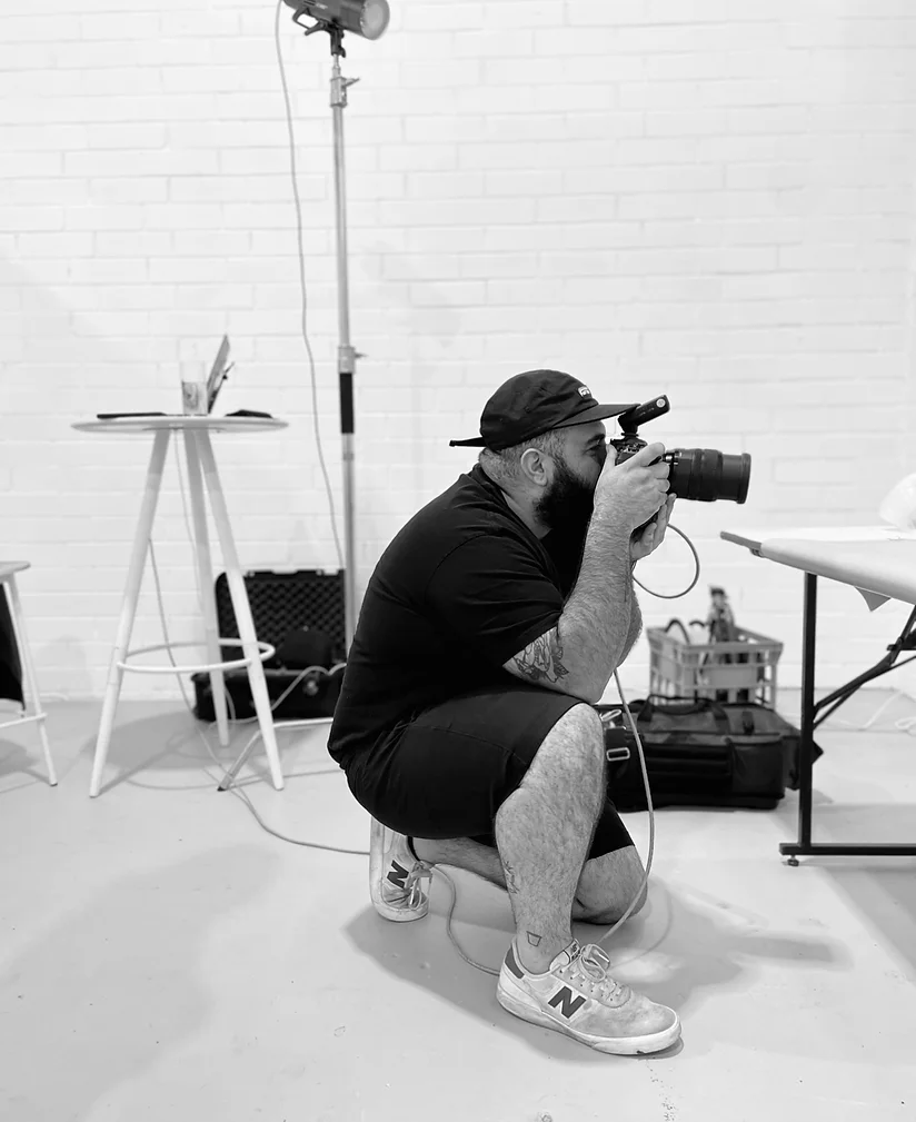 Crouching photographer in a studio takes a photo with a digital camera. Black and white view with photo equipment and a white brick wall.