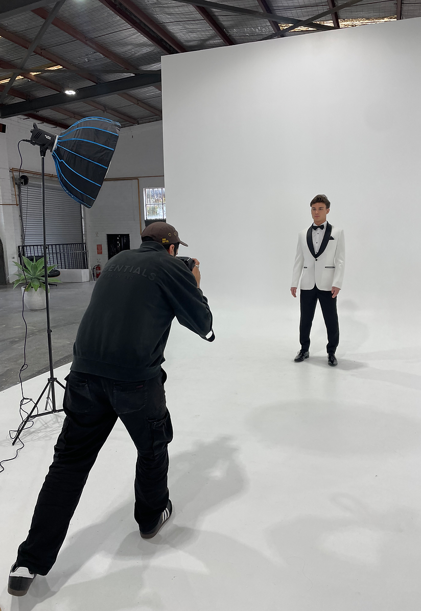 Man in a white tuxedo posing for a photographer in a photo studio with a white backdrop and studio lighting equipment visible.