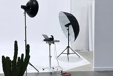 Photo studio setup with lighting equipment, a laptop on a rolling cart, and a potted cactus in a bright, minimalistic indoor space.