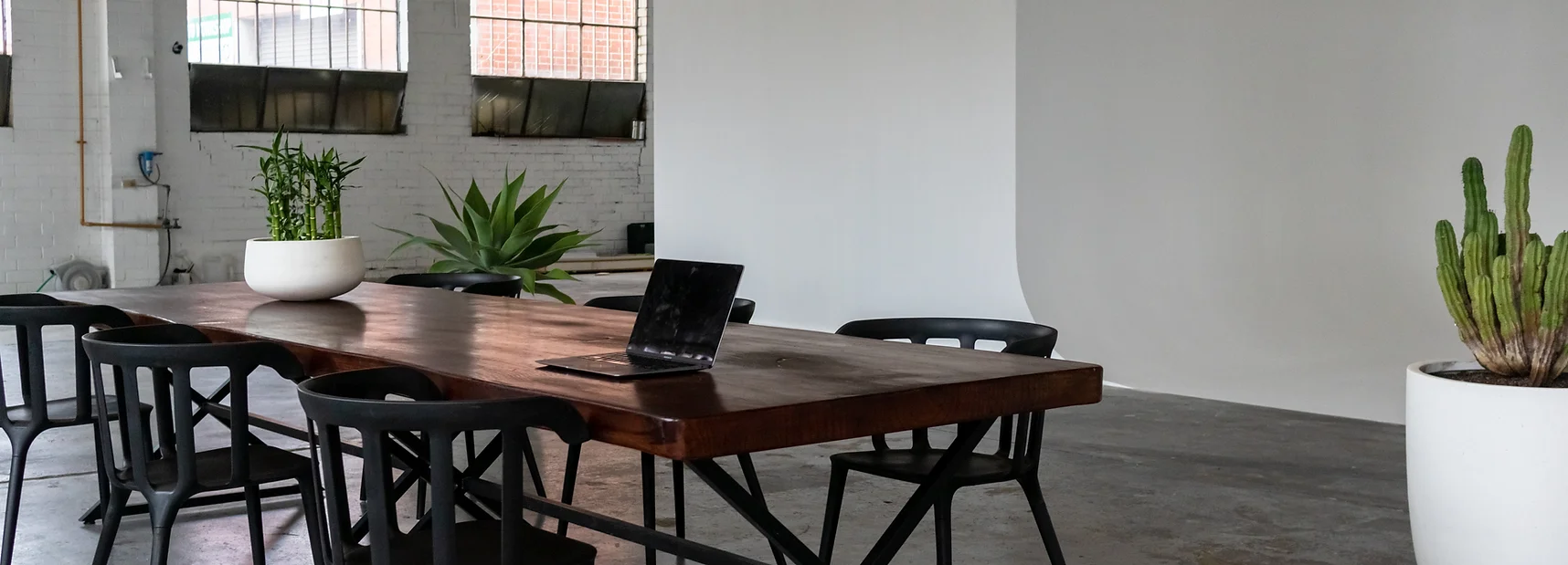 Conference room with a wooden table, black chairs, laptop, and plants in a white studio with large windows and natural light.