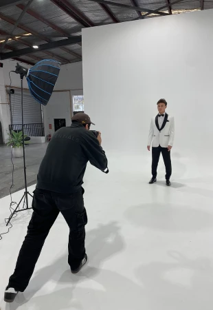 Photographer in black jacket and dark cap capturing a man in a white tuxedo jacket on a seamless white studio backdrop.