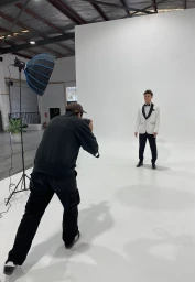 Photographer in black jacket and dark cap capturing a man in a white tuxedo jacket on a seamless white studio backdrop.